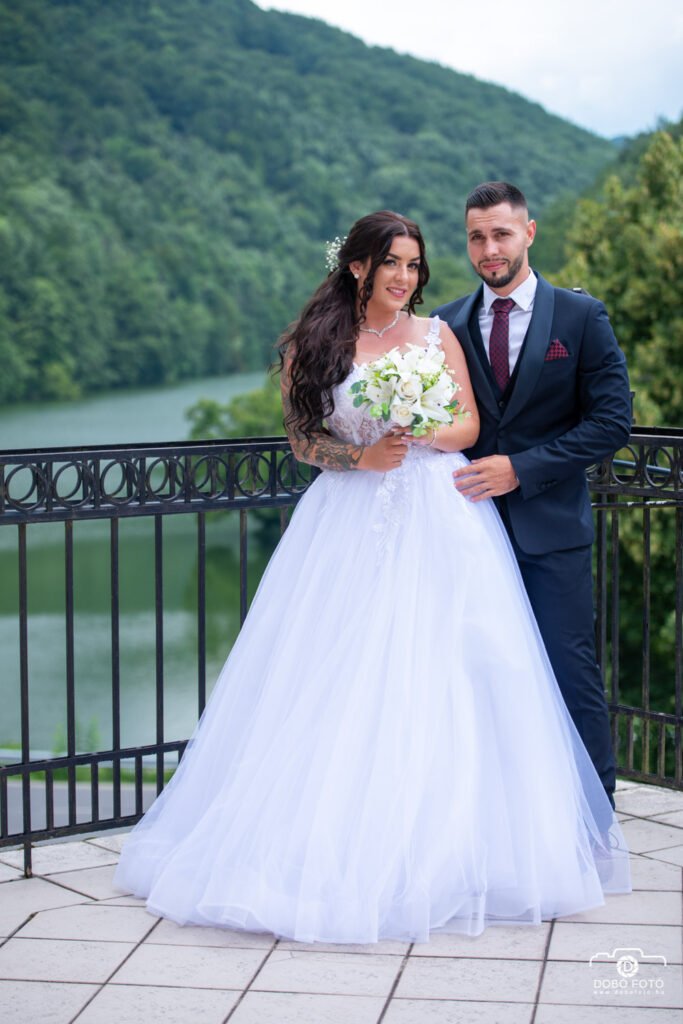Esküvői pár a tó felett, természetes környezetben. Menyasszony fehér, hercegnős ruhában, vőlegény sötétkék öltönyben. Profi esküvői fotózás, természetes fények, romantikus hangulat, professzionális esküvői fotós munka.