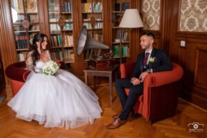 menyasszony és a vőlegény stílusos könyvtárszobában ülnek, piros fotelokban. A menyasszony csodás fehér ruhájában, virágcsokorral a kezében mosolyog, míg a vőlegény elegáns öltönyben figyelmesen néz rá. A háttérben klasszikus könyvespolcok és egy antik gramofon látható, amelyek különleges atmoszférát teremtenek.