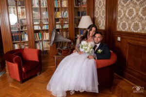 Kreatív fotózás. Az ifjú pár egy elegáns könyvtárszoba hangulatos foteljében ül, miközben egymásra mosolyognak. A menyasszony gyönyörű fehér ruhát visel, kezében virágcsokorral, a vőlegény pedig stílusos öltönyben. A háttérben könyvespolcok, antik gramofon és klasszikus berendezés látható, ami különleges, romantikus atmoszférát teremt.