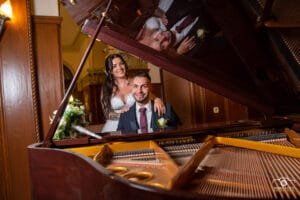 Az ifjú pár egy elegáns zongoránál látható, ahol a vőlegény öltönyben ül, a menyasszony pedig gyönyörű fehér ruhájában áll mögötte, és kezeit a vállára helyezi. A zongora fényes fedelén mindkettőjük tükörképe megjelenik, amely romantikus és egyedi hangulatot ad a képnek. A háttérben elegáns, klasszikus belső tér látható.