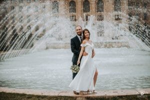 Esküvői fotózás a Debreceni Egyetem szökőkútjánál, a pár elegáns ruhában áll a vízpermet előtt, a menyasszony kezében virágcsokor.