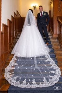 Az ifjú pár egy elegáns, fa burkolatú lépcsőházban áll, ahol a menyasszony gyönyörű, hímzett uszályos ruhája lágyan terül el a lépcsőkön. A vőlegény kifinomult öltönyben mosolyogva nézi kedvesét. A romantikus fények és a klasszikus belső tér kiemelik az esküvő meghitt pillanatait.