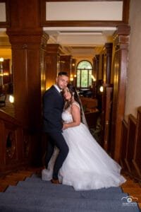 Az ifjú pár egy történelmi stílusú épület elegáns fa berendezései között áll, a háttérben nagy íves ablakok és lágy fények teremtenek romantikus hangulatot. A menyasszony gyönyörű fehér ruhájában, a vőlegény klasszikus öltözetben mosolyognak egymásra, kiemelve az esküvő eleganciáját és meghittségét.