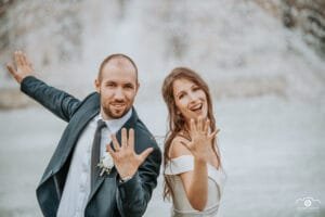 Vidám esküvői pár szökőkút előtt pózol, büszkén mutatva jegygyűrűjüket, kreatív esküvői fotózás.
