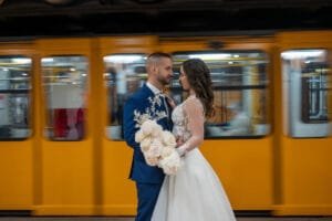 Romantikus esküvői portré sárga metrószerelvény előtt, csipkés hosszúujjú menyasszonyi ruha, kék öltöny, fehér rózsacsokor, mozgásban lévő háttér.