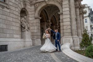 Táncos mozdulat az Operaház árkádjai alatt, forgó menyasszonyi ruha csipke felsőrésszel, kék öltönyös vőlegény, fehér csokor, budapesti kreatív esküvőfotózás.