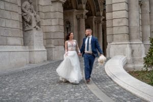 Spontán esküvői fotó az Opera árkádsora alatt, vidám ifjú pár sétál kézenfogva, csipkés-tülles menyasszonyi ruha, kék öltöny, fehér csokor.