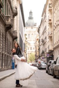 Emelős kreatív fotó szűk budapesti utcán, háttérben a Bazilika kupolája, menyasszony fehér tüllszoknyában és fekete bőrdzsekiben, vőlegény kék öltönyben.