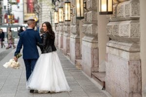 Hátulról fotózott ifjú pár sétál, fehér menyasszonyi ruha fekete bőrdzsekivel, kék öltöny, fehér csokor, Andrássy úti oszlopos épület előtt.