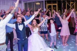 Esküvői party csúcspontja: ifjú pár és vendégek YMCA táncmozdulat közben, LED táncparketten. Fehér menyasszonyi ruha, kék mellény, rózsaszín-lila megvilágítás, professzionális rendezvényfotózás Debrecenben.