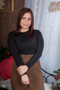 Karácsonyi portréfotózás Debrecenben, elegáns fekete felsőben és barna nadrágban mosolygó hölgy, ünnepi fényekkel a háttérben.