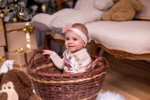 Mosolygó kisbaba fonott kosárban, karácsonyi díszítéssel és ajándékokkal a háttérben, megörökítve Debrecen hangulatos fotóstúdiójában.