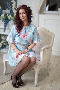 Egy nő világoskék, virágmintás ruhában ül egy elegáns, klasszikus stílusú karosszékben. A háttérben virágdíszek és világos enteriőr látható, melyek a kép barátságos és kifinomult hangulatát hangsúlyozzák.