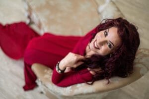 Egy nő elegáns vörös ruhában fekszik egy díszes kárpitozású kanapén, mosolyogva nézve a kamerába. A fotó letisztult és elegáns enteriőrben készült, kiemelve a nőiességet és a könnyedséget.