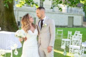 Esküvői fotózás. Az ifjú pár egymásra mosolyogva áll a szabadtéri szertartás helyszínén, háttérben fehér széksorok és üvegvázás dekoráció.