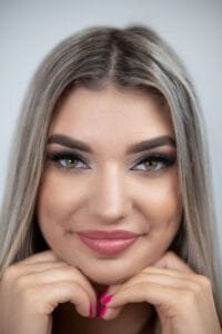 Portré fotó - Dobófotó stúdió. Egy közeli portré egy fiatal nőről, amely az arca részleteit hangsúlyozza. A smink finoman kiemeli a szemeit és ajkait, a természetes hatású smink tökéletes összhangban van a ragyogó mosollyal. A világos háttér a modell vonásaira fókuszál.