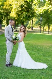 A menyasszony fehér csipkeruhában, kezében fehér virágcsokorral mosolyog, mellette vőlegénye világosszürke öltönyben a kastélypark napsütötte gyepén.
