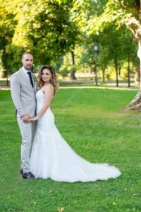 Az ifjú pár a kastélypark zöld gyepén, mögöttük lombos fák - a menyasszony hosszú uszályos csipkeruhában, a vőlegény világosszürke öltönyben.