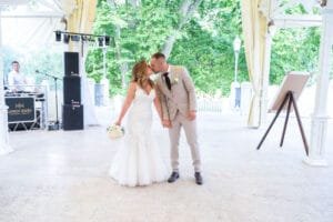 A menyasszony és vőlegény a téglási kastély teraszán csókolózik, háttérben DJ-pult és zöldellő fák.