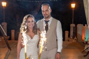 A menyasszony és vőlegény mosolyogva állnak az esti teraszon, előttük csillagszórók szikráznak, háttérben kandeláberek fénye világít.