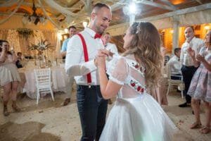 Az ifjú pár első tánca az átöltözés után - a menyasszony magyaros, piros rózsás fehér ruhában, a vőlegény piros nadrágtartóval, körülöttük táncoló vendégek a díszteremben.