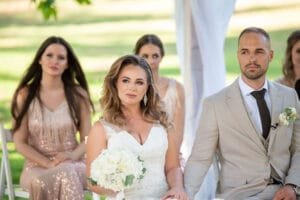 Menyasszony fehér csipkeruhában, fehér rózsacsokorral és vőlegény világosszürke öltönyben a szertartáson, mögöttük koszorúslány rózsaszín flitteres ruhában.