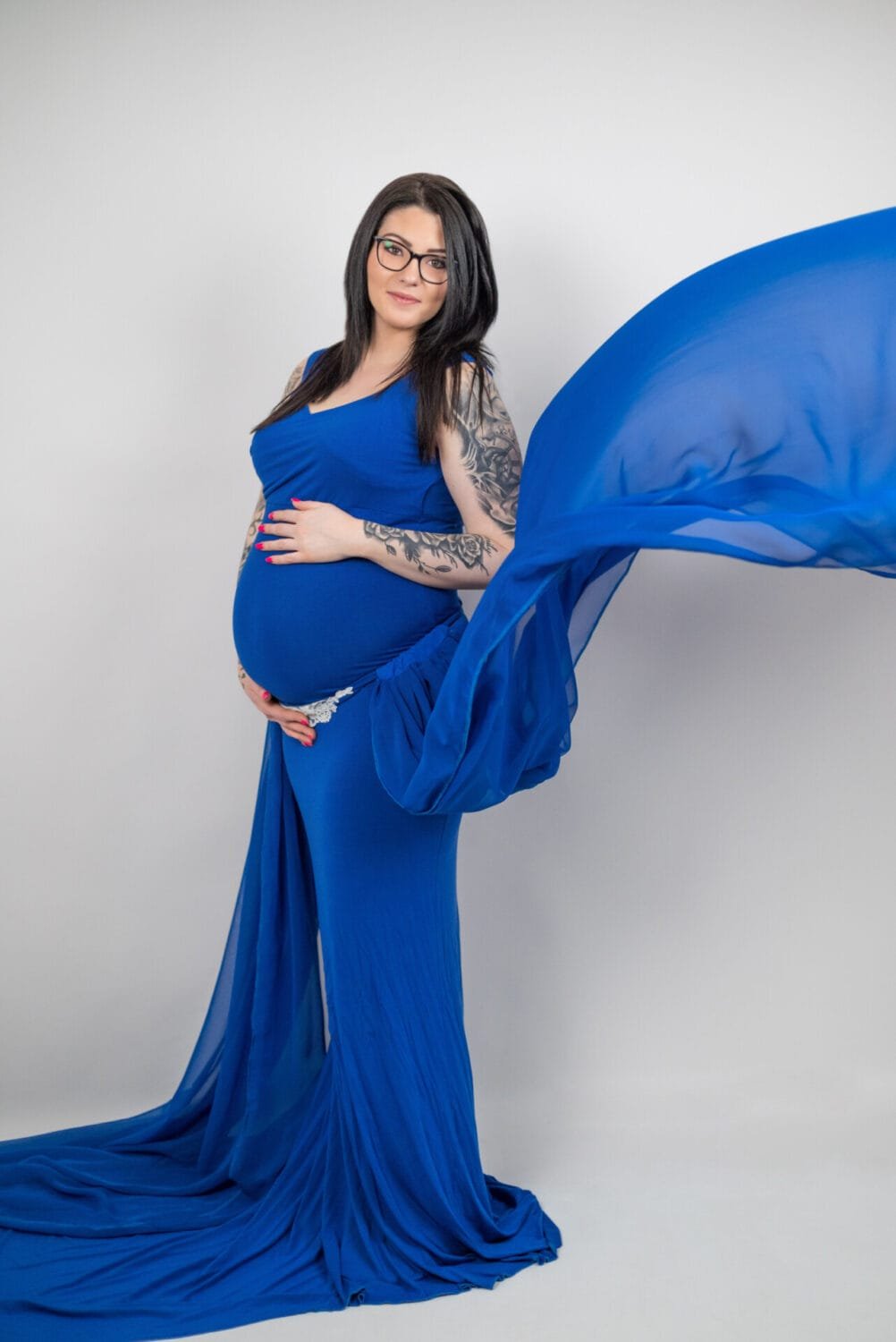 Kismama fotózás Debrecenben - Kismama kék, légies ruhában, gyönyörű pózban egy stúdióban. A ruha lágy szövete lendületesen terül el, miközben a kismama magabiztosan és boldogan mosolyog. A fotó elegáns és harmonikus hangulatot áraszt.