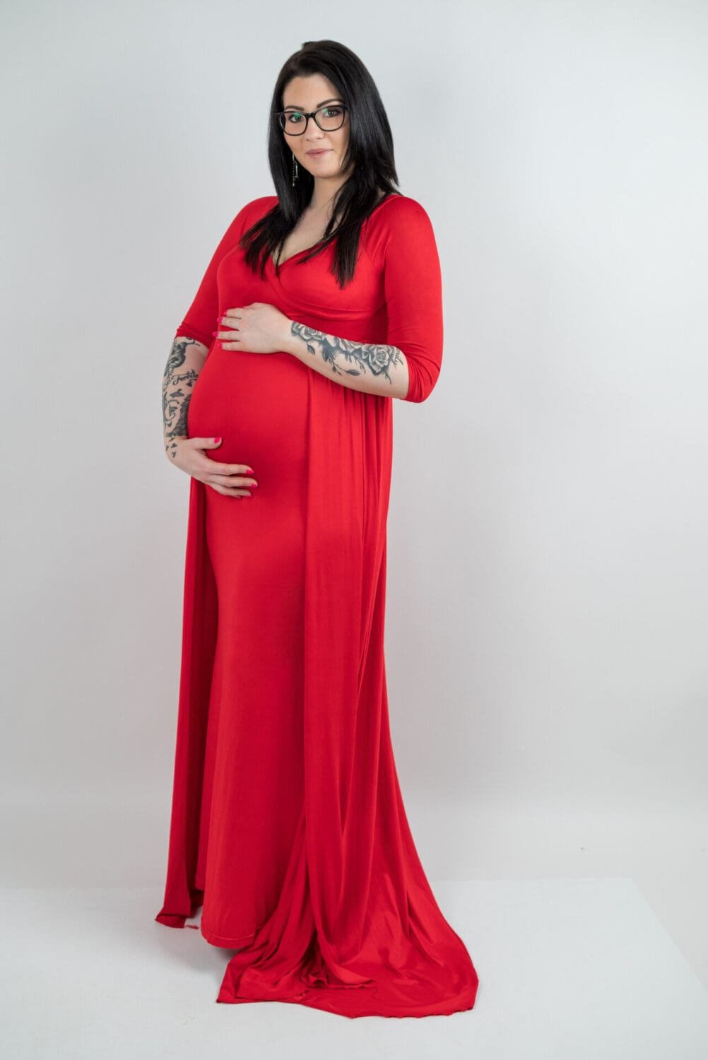 Pocakos fotózás során a kismama élénk piros, elegáns ruhában, kezét gyengéden a pocakjára helyezve mosolyog egy stúdióban készült fotón. Az egyszerű háttér kiemeli a ruha színét és a pillanat meghittségét.