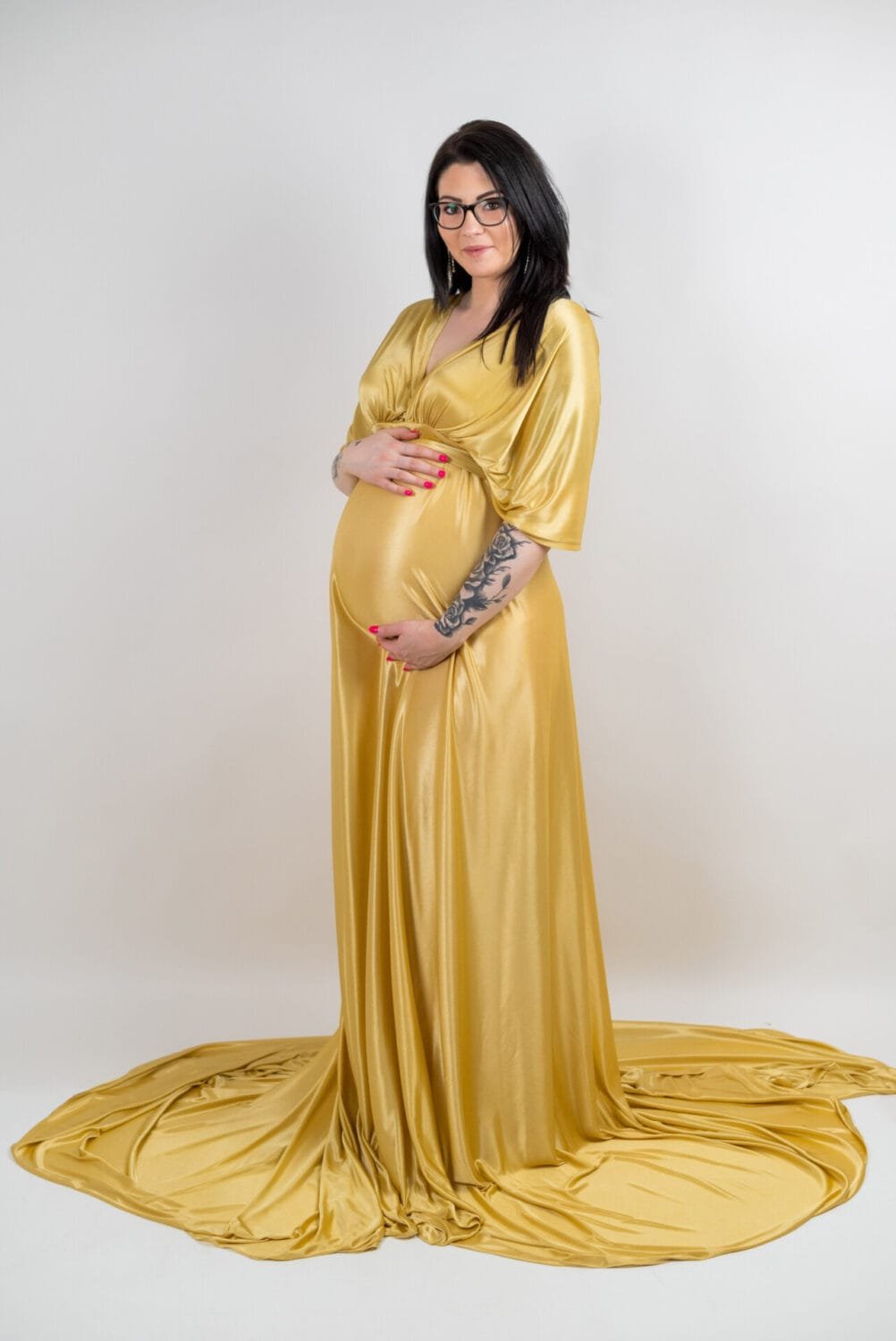 Kismama fotózás Debrecen - Ajándék kismama ruha fotózáshoz Debrecenben. Kismama arany színű, hosszú, elegáns ruhában áll egy stúdióban, kezét szeretettel a pocakjára helyezve. A ruha lágy anyaga és a visszafogott háttér harmonikus, meghitt hangulatot teremt.