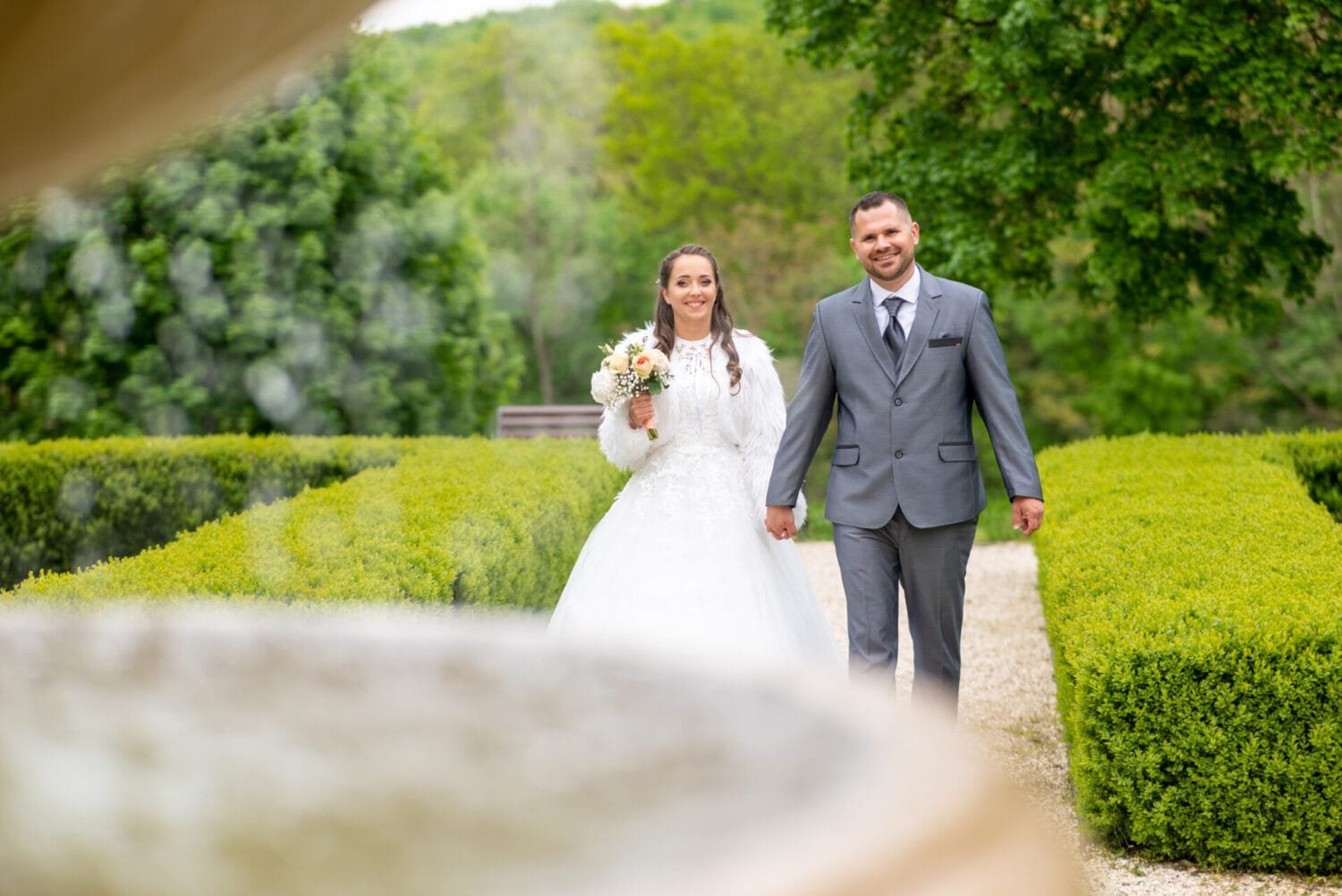 Ifjú pár kézen fogva sétál elegáns esküvői ruhában egy gyönyörű parkban, virágcsokorral a menyasszony kezében, romantikus pillanat a természetben.