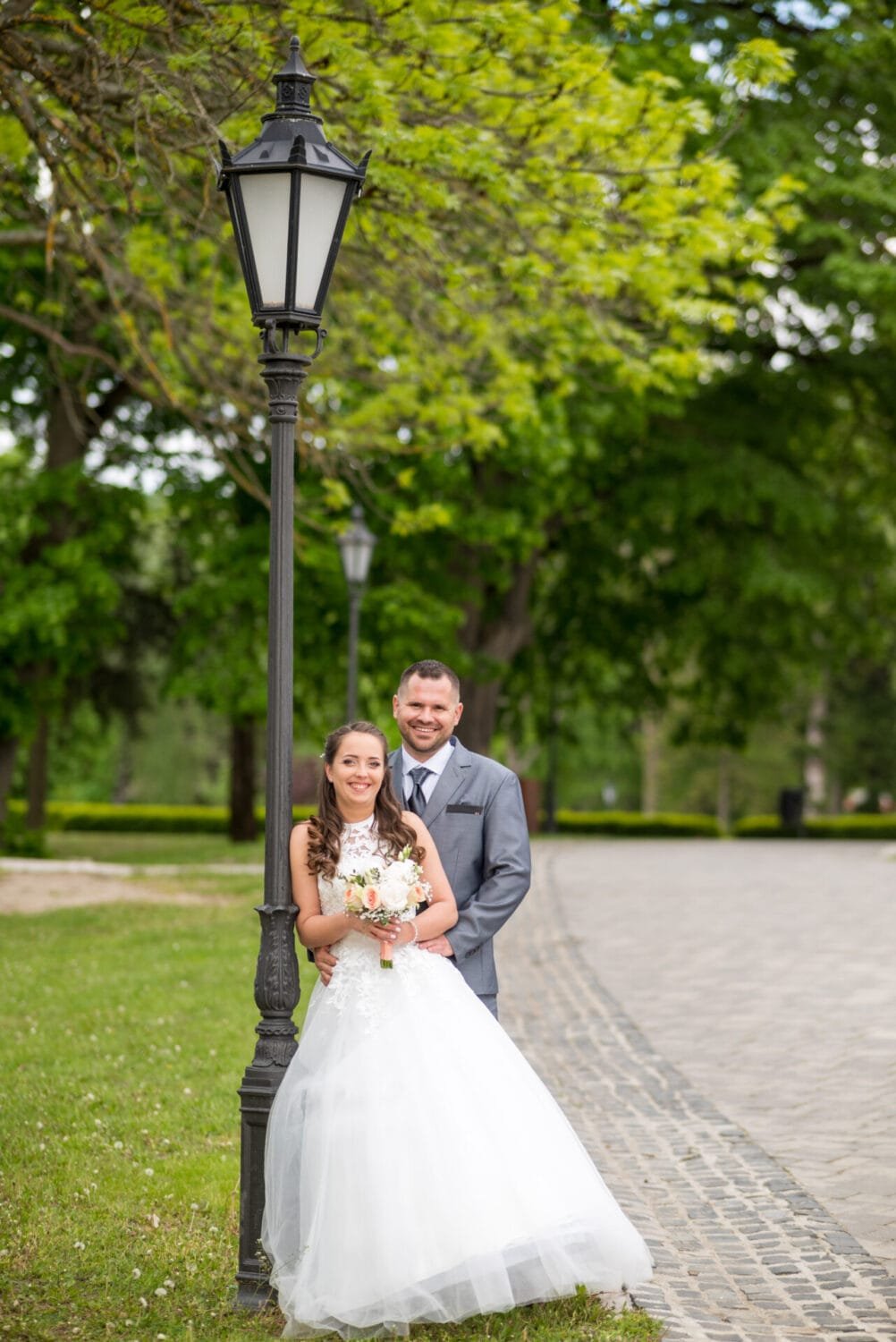 Menyasszony és vőlegény állnak egy hangulatos utcai lámpa mellett Szilvásvárad parkjában. A menyasszony hófehér ruhában, kezében virágcsokorral, míg a vőlegény elegáns öltönyben mosolyog. Körülöttük zöldellő fák és egy macskaköves sétány látható.
