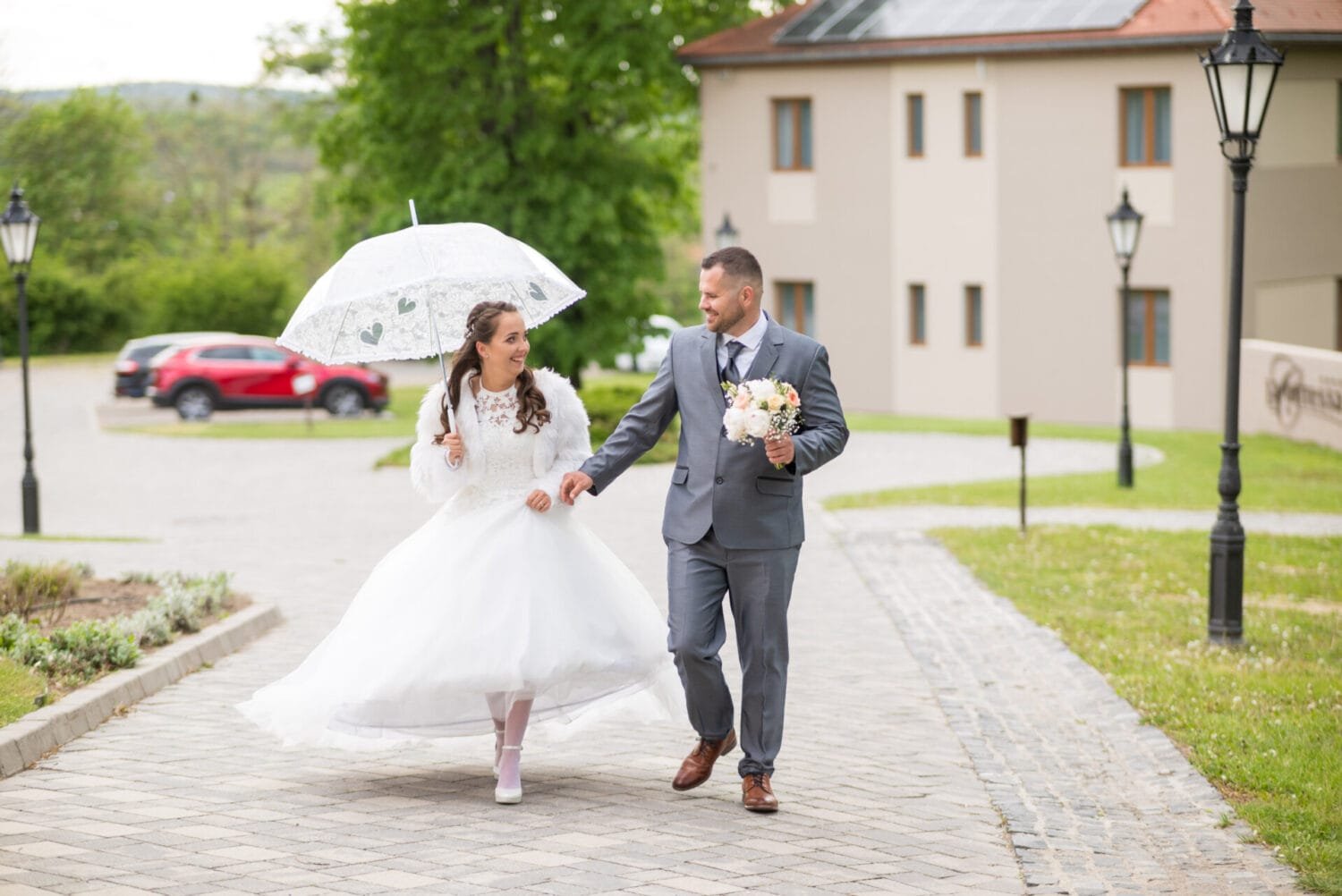 Menyasszony és vőlegény mosolyogva sétál egy romantikus helyszínen, ahol a menyasszony egy szív mintás áttetsző esernyőt tart, míg a vőlegény egy szép virágcsokrot. A háttérben modern épületek és természet látható.