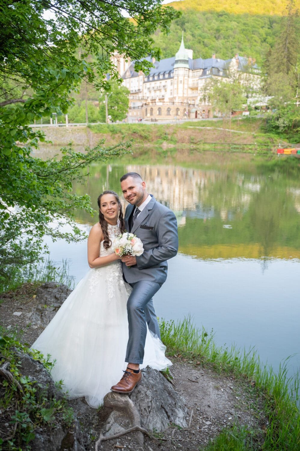 A menyasszony és a vőlegény a lillafüredi tó partján állnak, mögöttük a víz tükrében a híres Palotaszálló látható. A pár mosolyog, a menyasszony elegáns fehér ruhában tartja virágcsokrát, miközben a zöld növényzet és a tó idilli hátteret nyújt a képhez.