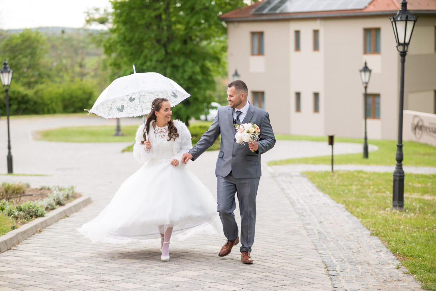 Esküvői fotós Debrecenben - A menyasszony és a vőlegény boldogan sétálnak kéz a kézben egy romantikus kastély udvarán. A menyasszony csipkés, szív alakú mintával díszített esernyőt tart, amely védi őket a könnyed esőtől, miközben a vőlegény gyönyörű csokrot tart a kezében. A háttérben a La Contessa kastély modern és klasszikus elemei ötvöződnek.