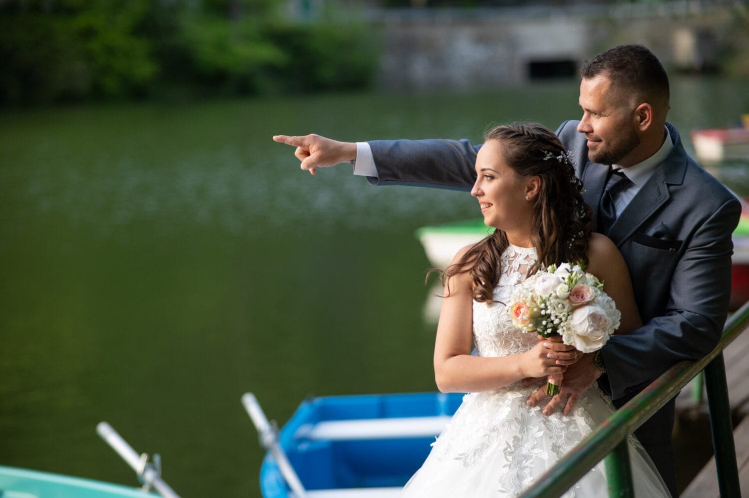 Egy romantikus pillanat Lillafüreden: a menyasszony és a vőlegény a tó partján állnak, a vőlegény mutat valamit a távolban, míg a menyasszony mosolyogva néz. A menyasszony kezében elegáns virágcsokor van, mögöttük csónakok és a zöld víz tükröződése látható.