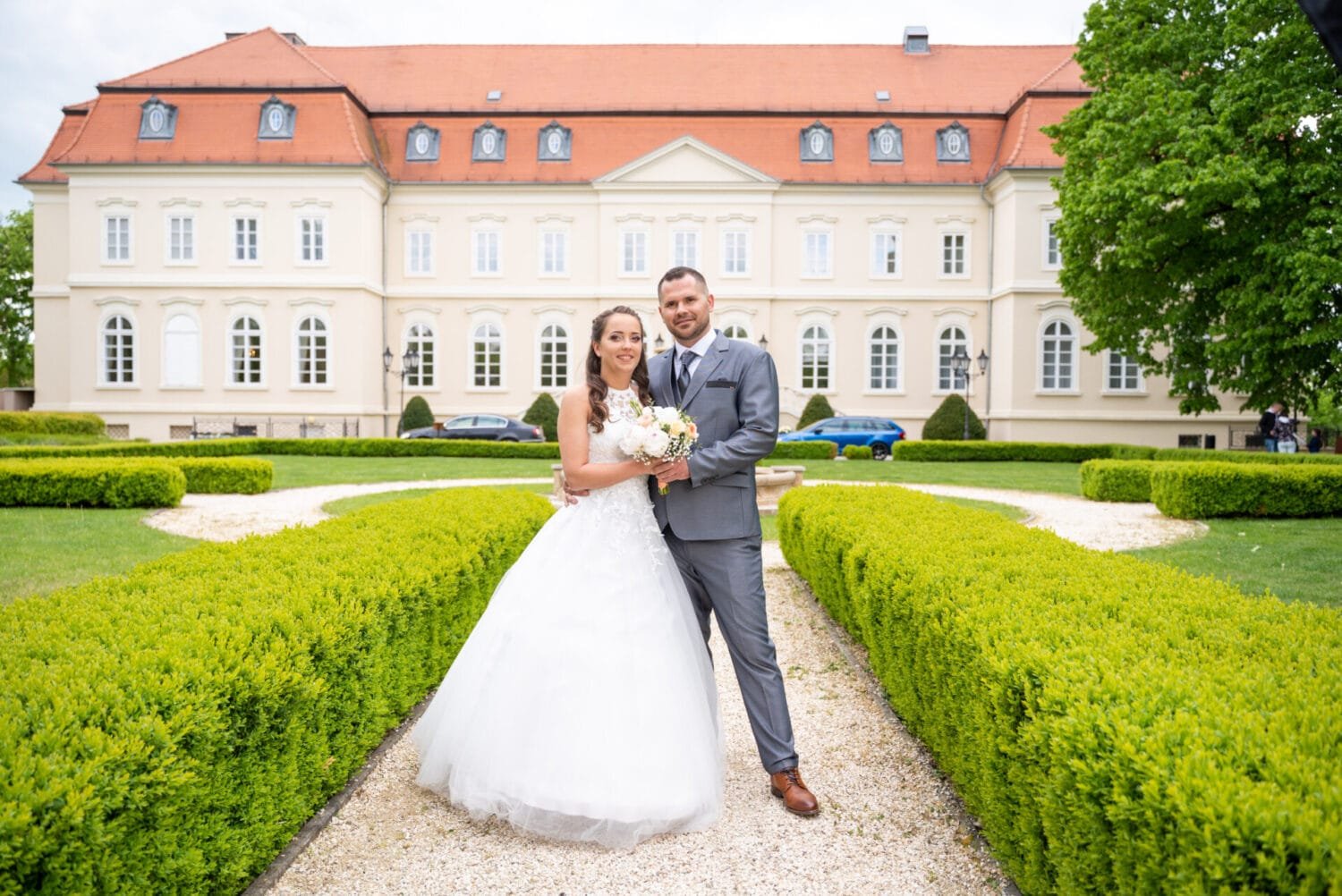 Ifjú pár esküvői ruhában egy gyönyörű kastély előtt, elegáns parkos környezetben készült romantikus esküvői fotón.