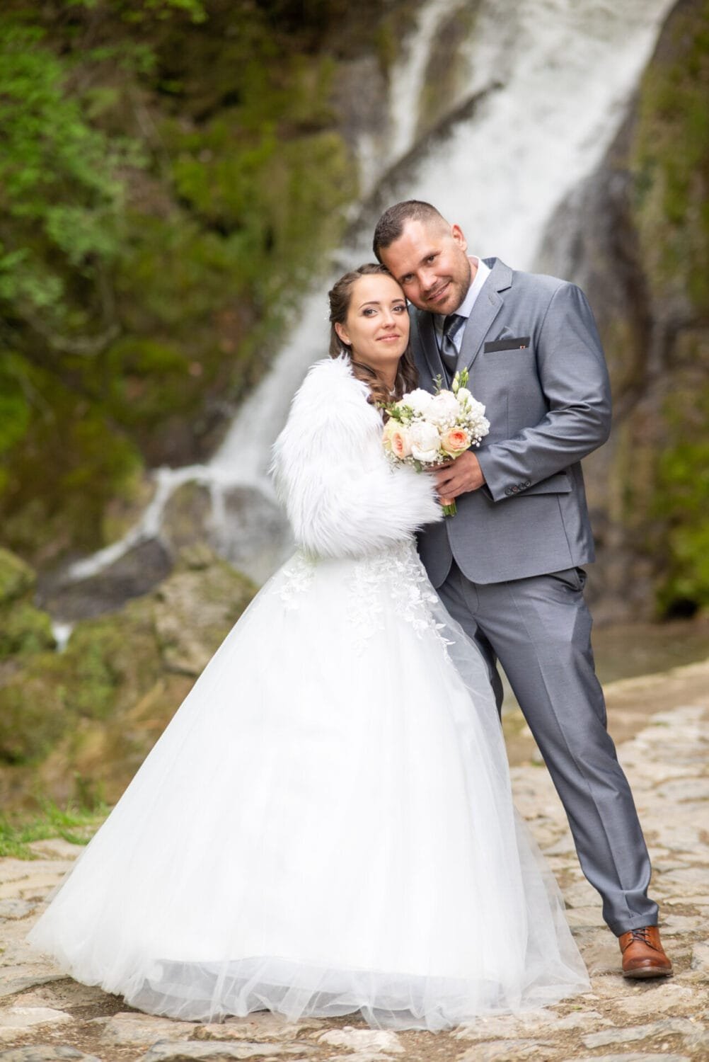 Egy boldog menyasszony és vőlegény szorosan egymáshoz bújik egy festői vízesés előtt. A menyasszony elegáns fehér ruhát és bolyhos fehér vállmelegítőt visel, kezében színpompás virágcsokorral, míg a vőlegény szürke öltönyben áll mellette. A háttérben a természet zöldje és a vízesés nyújt lenyűgöző díszletet.