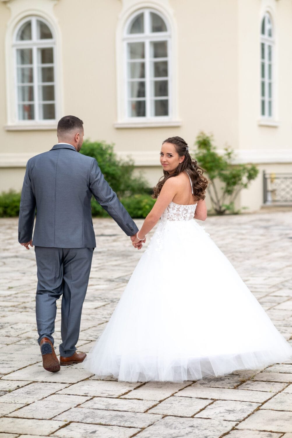 Egy romantikus pillanat, amikor a menyasszony és a vőlegény kézen fogva sétálnak a kastély udvarán. A menyasszony visszatekint a válla fölött, mosolyogva a kamerára, elegáns fehér ruhájában. A háttérben a kastély ablakai és zöld növényzet láthatóak.