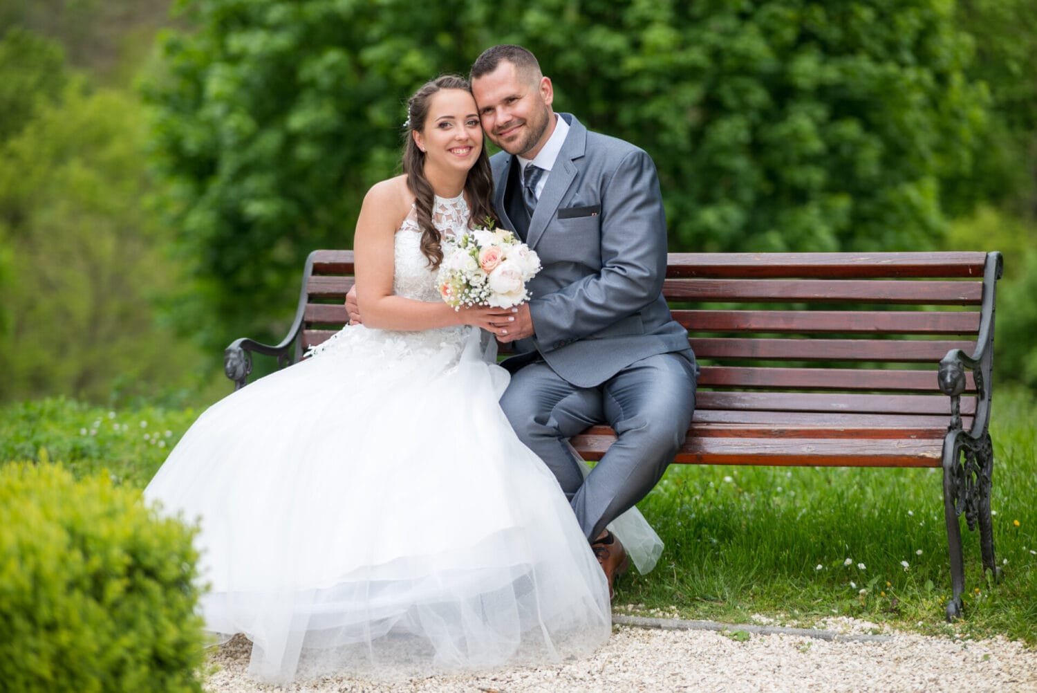 Menyasszony és vőlegény romantikus pillanatban, ahogy egy padon ülnek Lillafüred zöldellő környezetében. A menyasszony gyönyörű fehér ruhában és virágcsokorral, a vőlegény elegáns öltönyben.