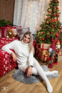 Fiatal nő fehér kötött ruhában karácsonyfa és ajándékok mellett, ünnepi hangulatú fotón, piros karácsonyi dekorációval körülvéve Debrecenben.