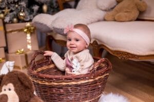 Mosolygó kislány fonott kosárban ülve karácsonyi háttérrel és ajándékdobozokkal körülvéve, ünnepi hangulatú fotón Debrecenben.