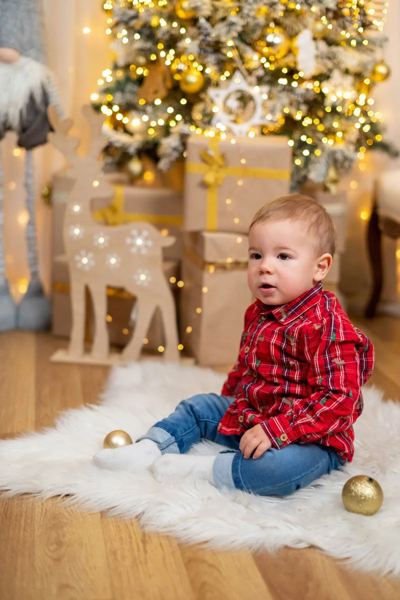 Egy kisfiú piros kockás ingben játszik a karácsonyi díszekkel, miközben a karácsonyfa és az ajándékok díszítik a hátteret. A kisfiú a földön ül, és boldogan nézi a körülötte lévő ünnepi dekorációkat.