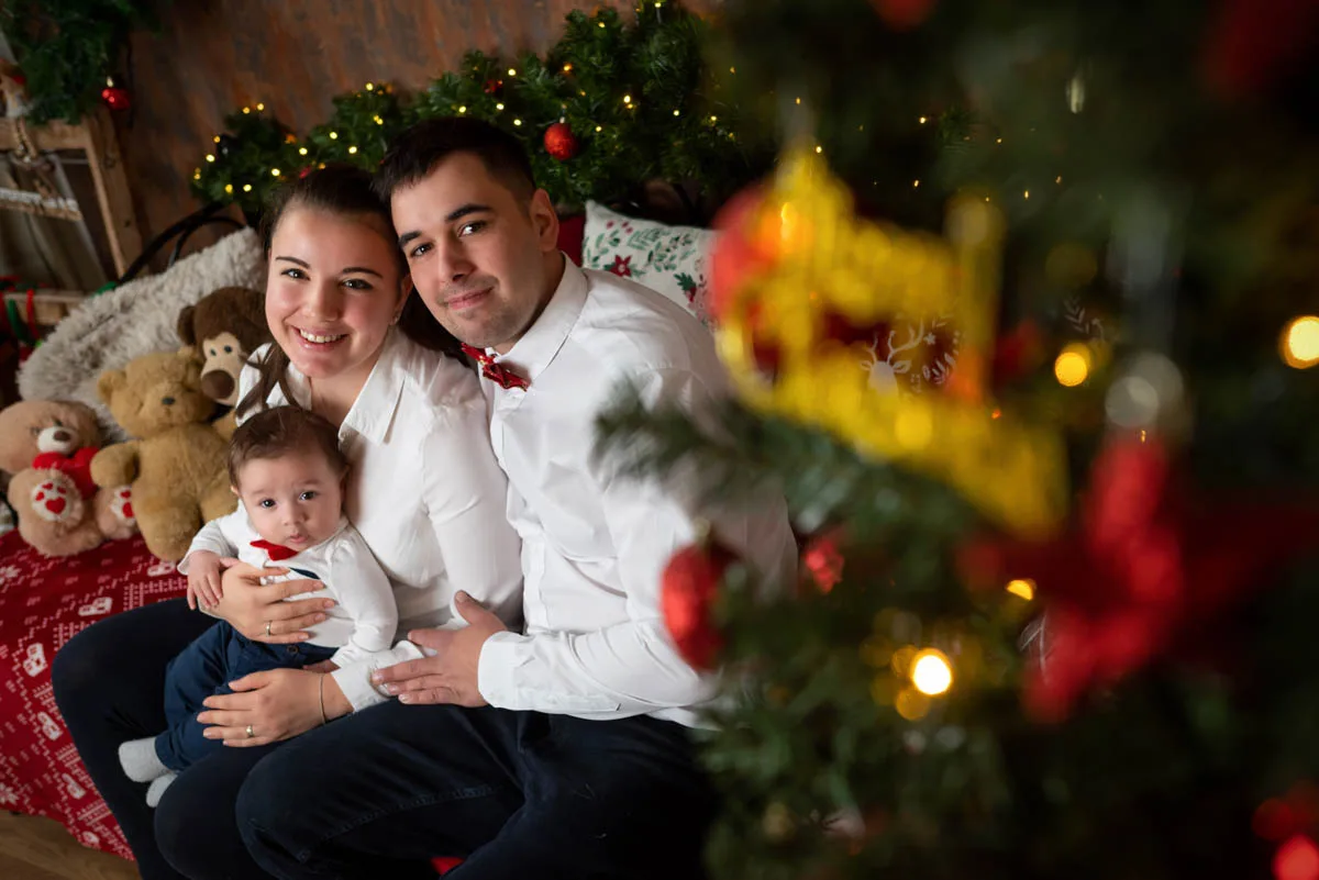 Egy boldog család karácsonyi hangulatban, fehér ingben és piros masniban. A kisfiú az anyukájával és apukájával pózol a fenyőfa mellett, miközben a háttérben karácsonyi díszek és plüss mackók láthatók.