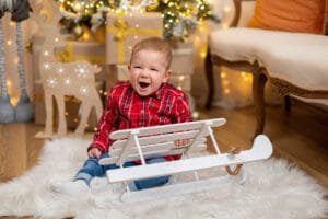 Egy kisfiú boldogan mosolyogva ül a fehér szánkón, miközben a karácsonyi díszek és a fenyőfa körül fények ragyognak. A család karácsonyi hangulata Debrecenben.