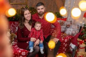 Egy fiatal család közös karácsonyi fotózáson, ünnepi díszletekkel, piros ruhákban, meghitt és örömteli hangulat Debrecenben.