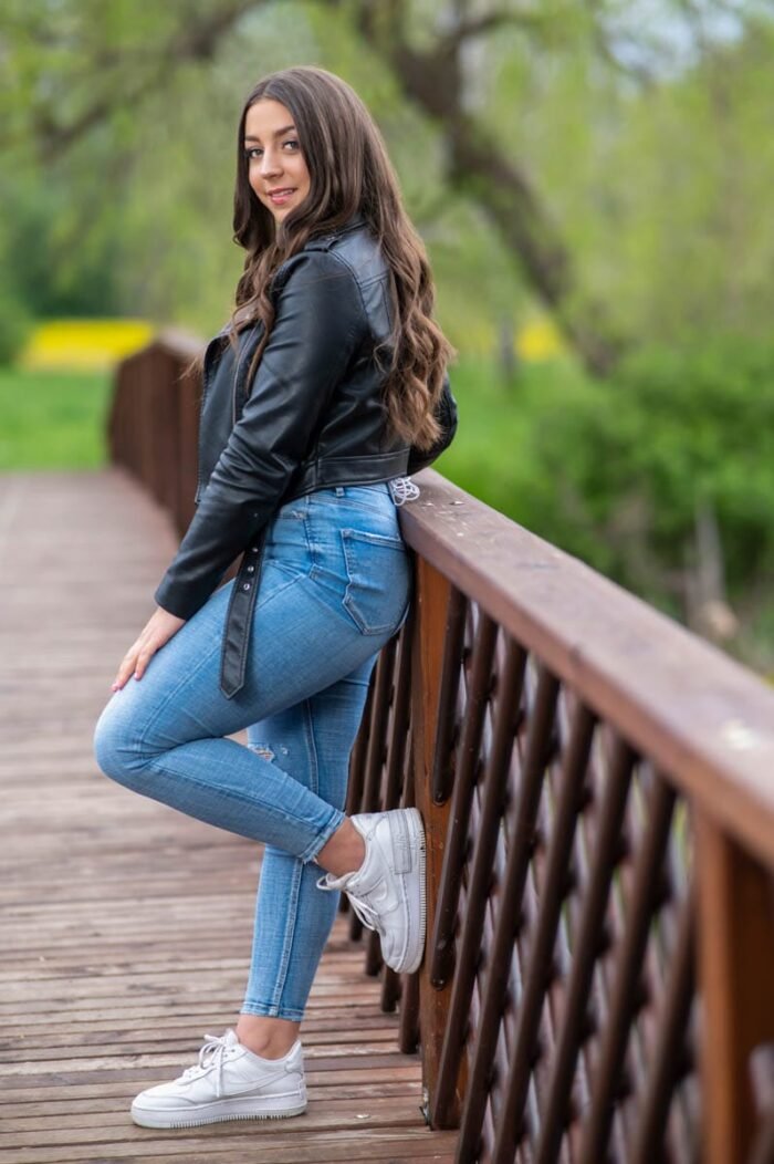 Fiatal nő fekete bőrdzsekiben és kék farmerben, fehér sportcipőben áll egy fa híd korlátjánál, mosolyogva, Debrecen közelében, a Vekeri-tónál.