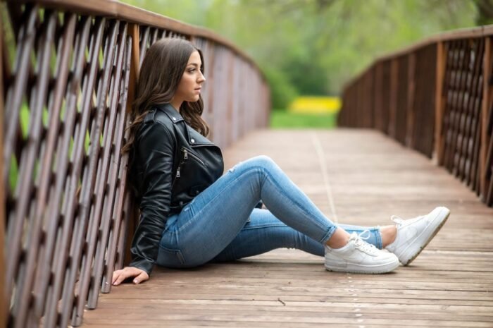 Fiatal nő fekete bőrdzsekiben és kék farmerben ül egy fa hídon, maga elé meredve, a háttérben tavaszi zöld és sárga táj látható.