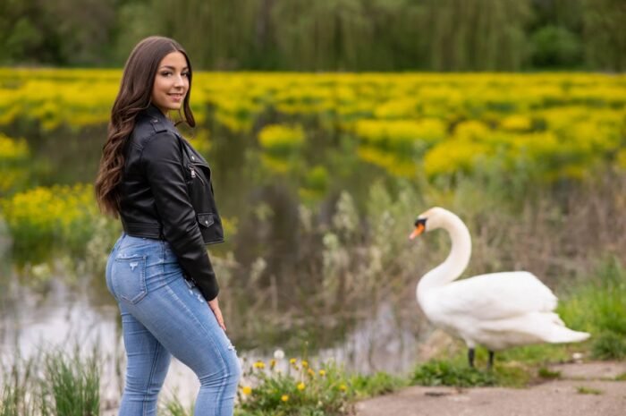 Fiatal nő fekete bőrdzsekiben és farmernadrágban mosolyogva pózol a Vekeri-tónál, egy hattyú és sárga virágokkal teli tájjal a háttérben, Debrecen közelében készült portréfotó.