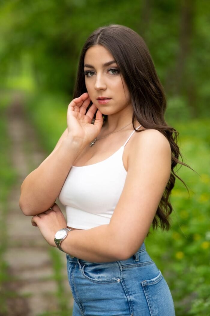 Fiatal nő fehér topban és farmernadrágban, zöld erdei háttér előtt pózol, természetes fényekkel készült portréfotó.