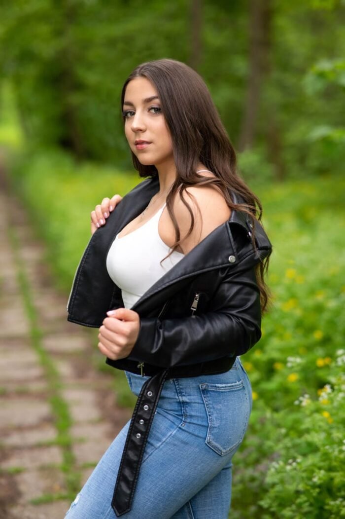 Fiatal nő fekete bőrdzsekiben és farmernadrágban pózol egy zöld, erdős háttér előtt, természetes fényben készült portréfotó.